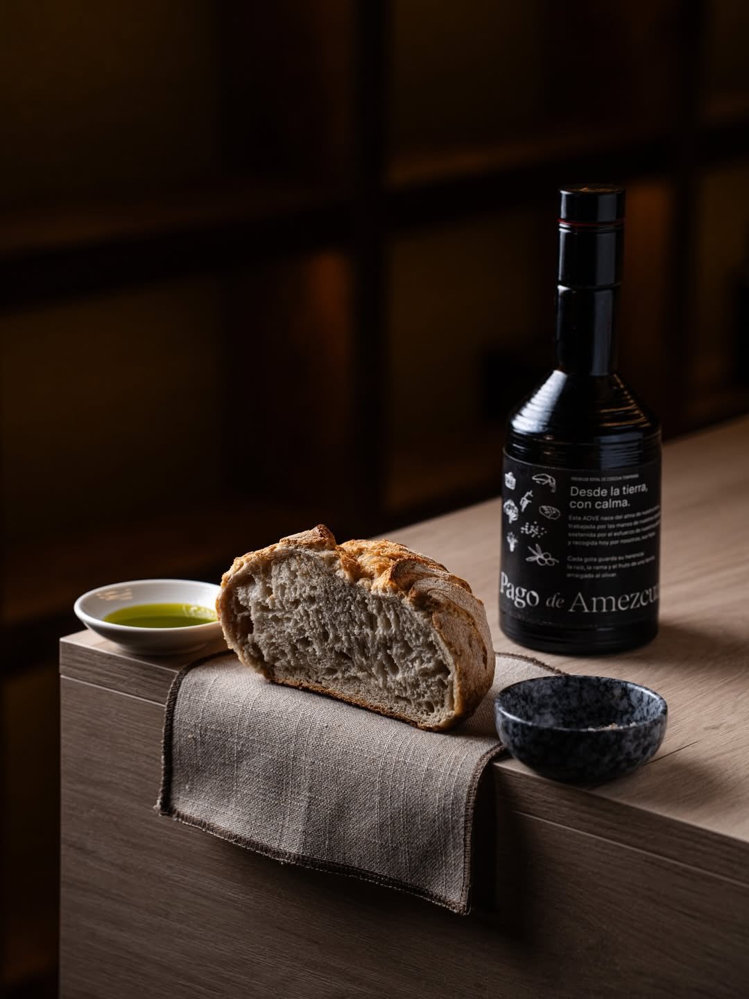 Botella verde de AOVE Pago de Amezcua sobre mesa Gourmet con pan y aceite