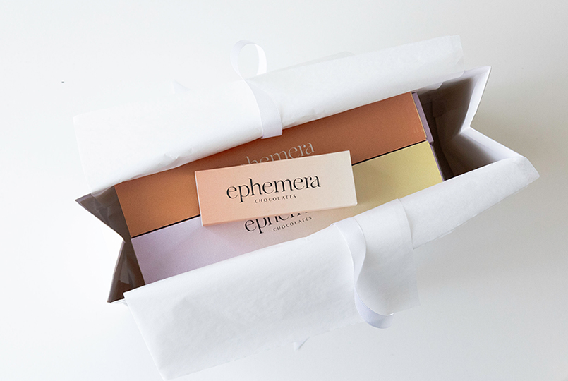 Caja de chocolates Ephemera envuelta en papel de seda dentro de una bolsa de papel blanca con packaging elegante en colores pastel.