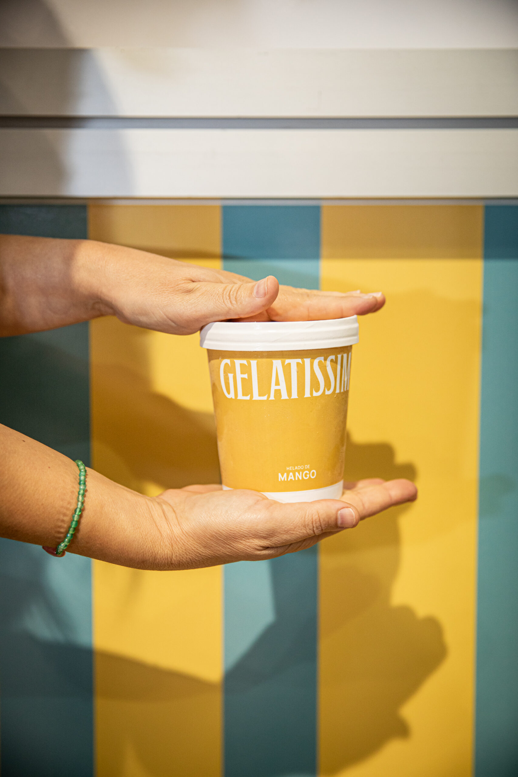 Persona sujetando tarrina de helado de mango Gelatissim sobre fondo de rayas.