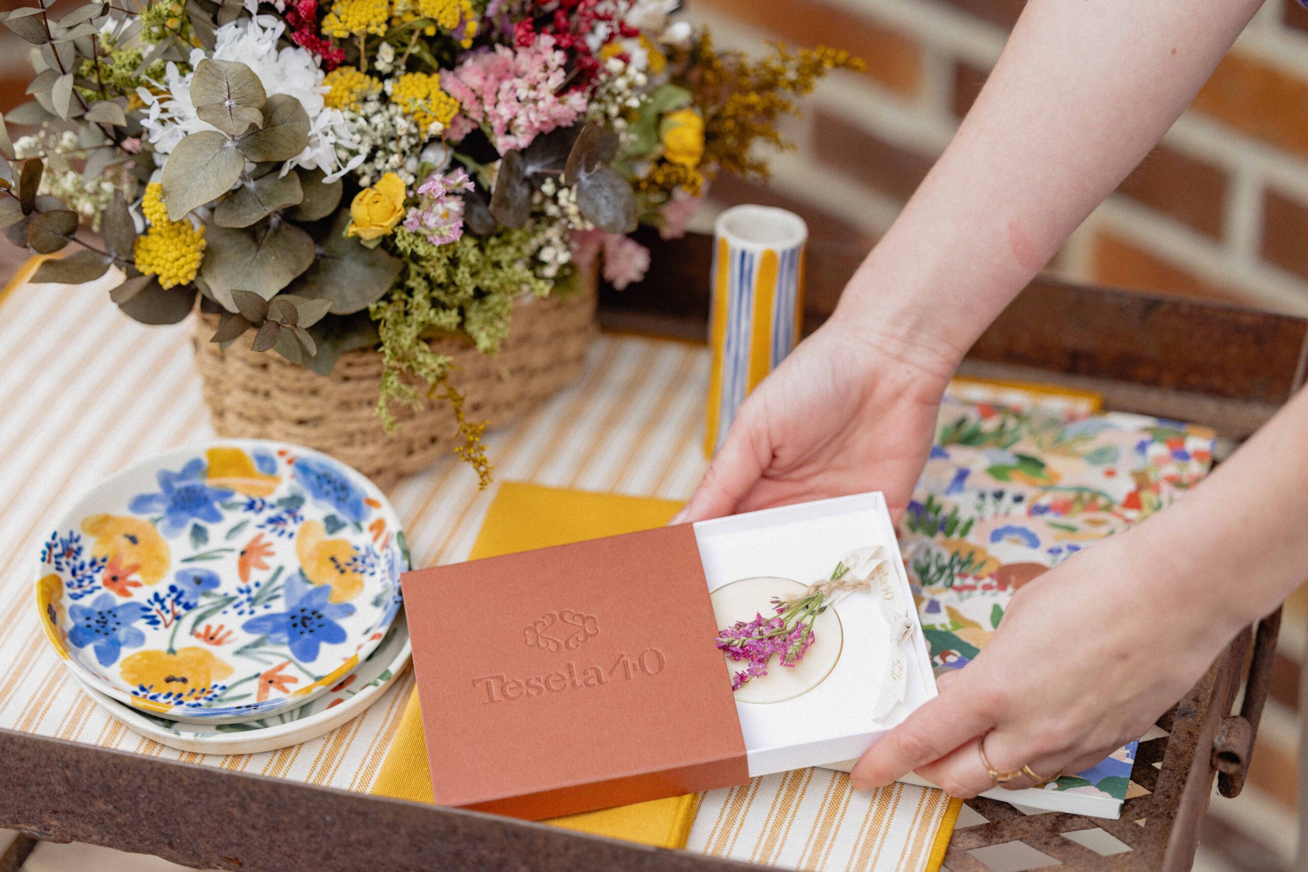 Presentación de producto: estuche de marca Tesela 40 en tono terracota junto a vajilla ilustrada y ramo de flores secas en un entorno artesanal.