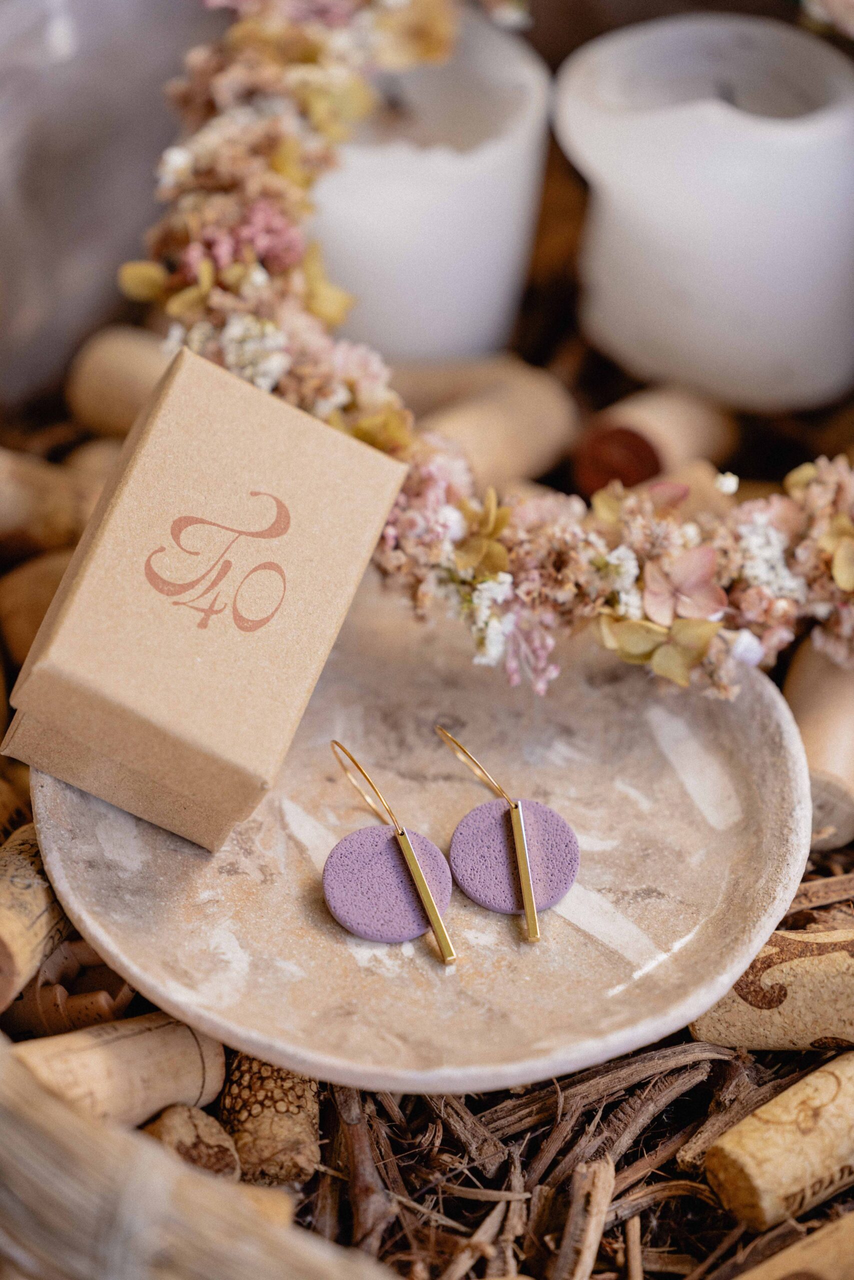 Diseño de packaging para joyería: caja de cartón kraft con logotipo estampado y pendientes de cerámica morada sobre plato de piedra decorativo.