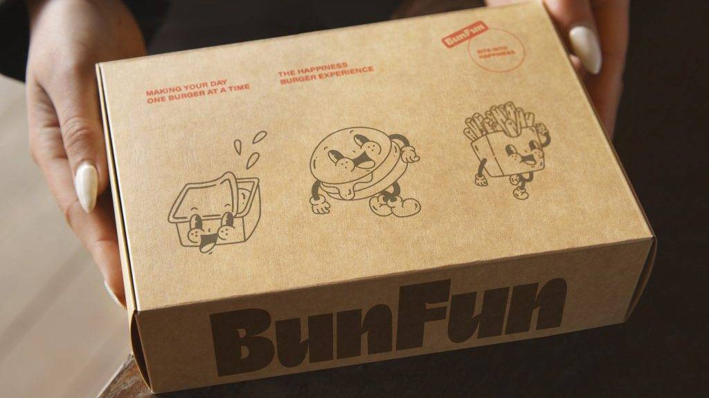 Diseño de packaging para hamburguesas: manos sosteniendo una caja de cartón kraft personalizada con ilustraciones de personajes cartoon (salsa, hamburguesa y patatas fritas) y el logotipo de BunFun.