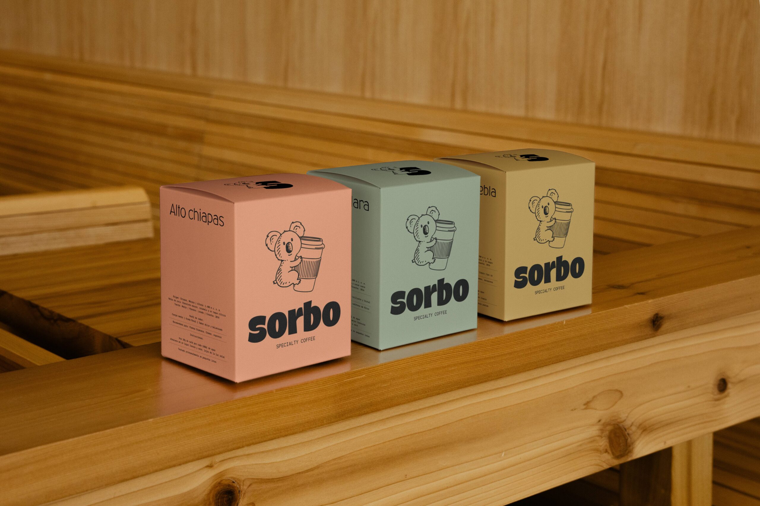 Diseño de packaging para café: tres cajas cúbicas en colores salmón, verde agua y mostaza alineadas sobre una superficie de madera.