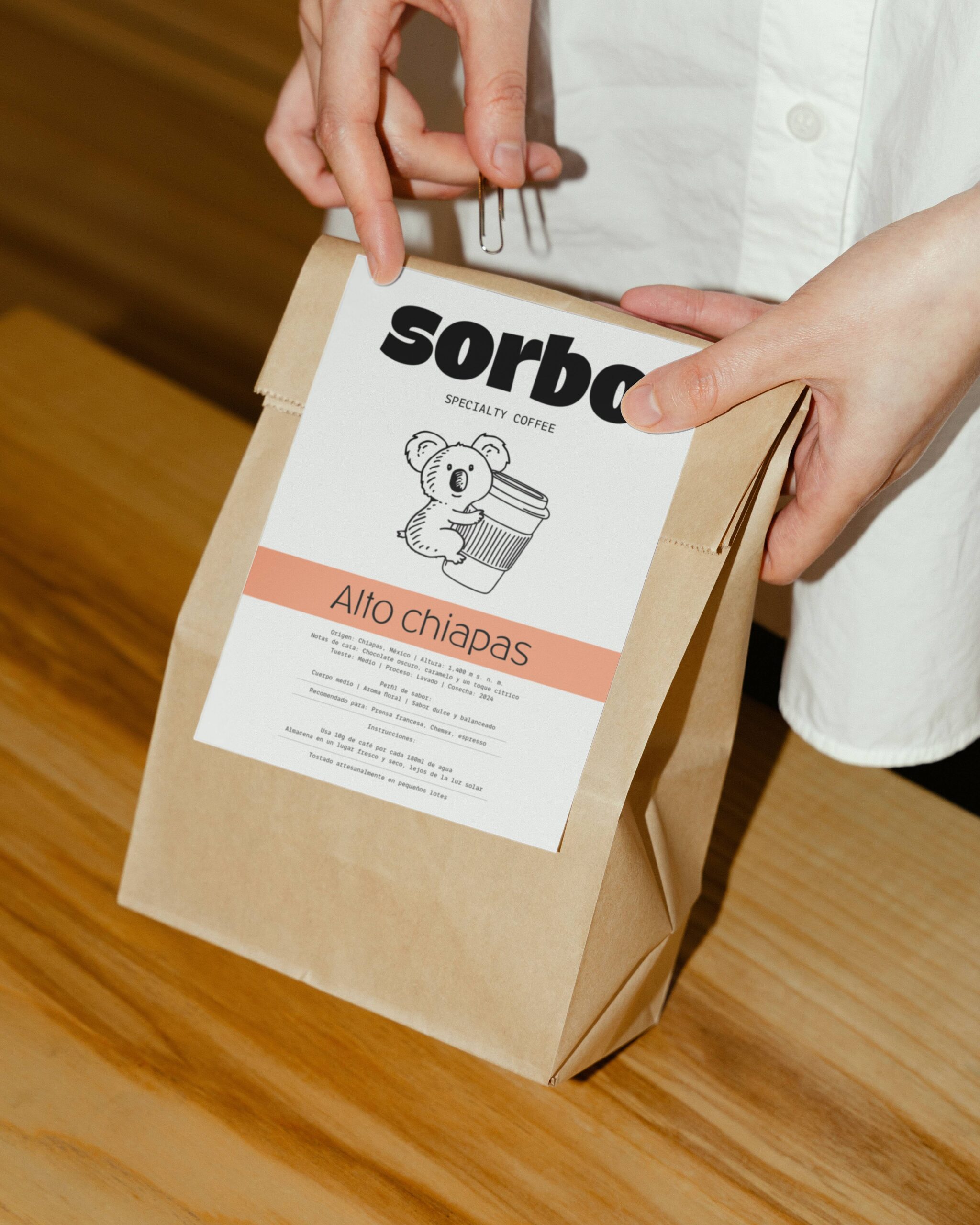 Detalle de packaging artesanal: manos cerrando una bolsa de café de papel kraft con un clip metálico y una etiqueta informativa de "Alto Chiapas".