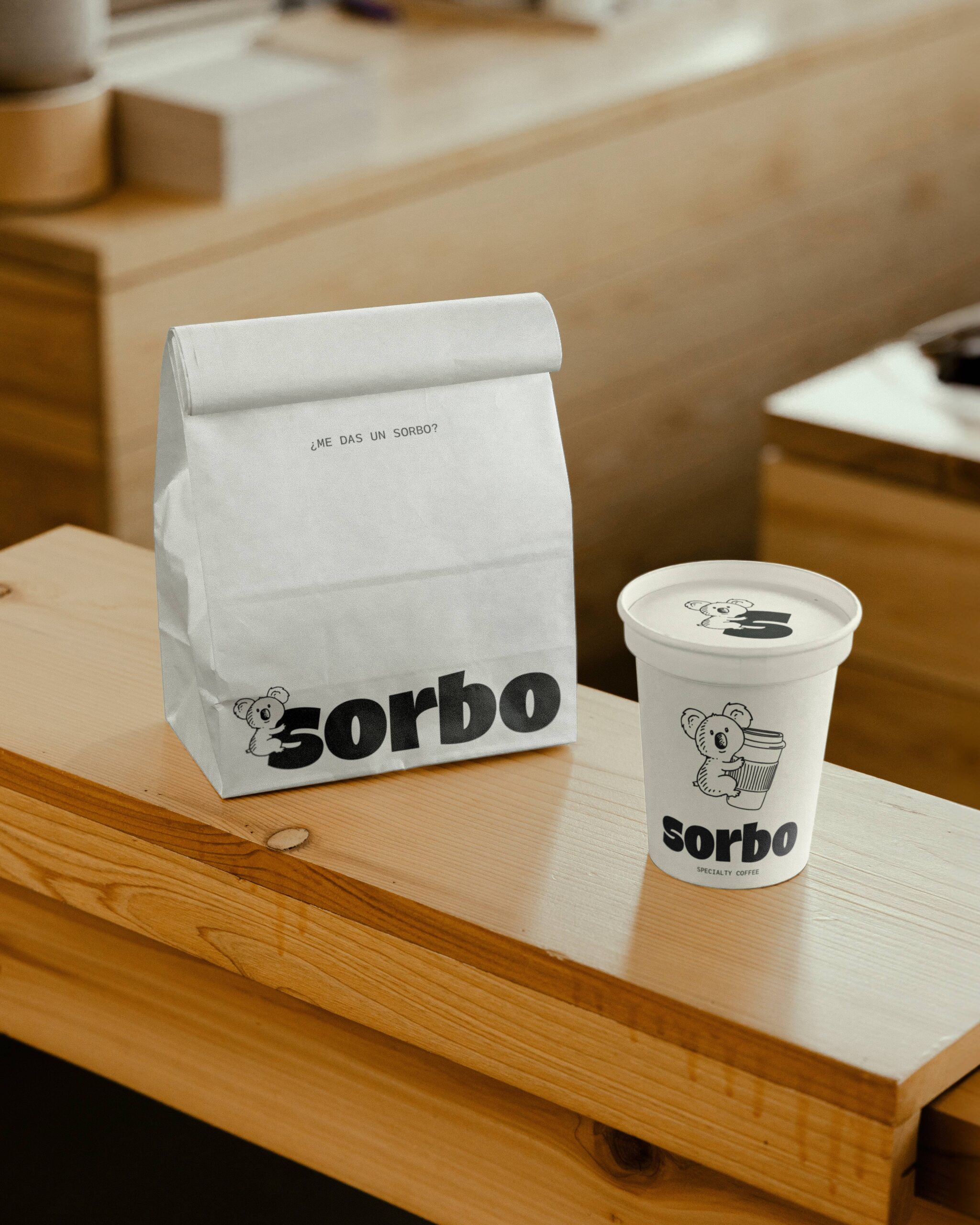 Pack de takeaway de Sorbo: bolsa de papel blanca con mensaje "Me das un sorbo?" junto a un vaso de café para llevar con ilustración de koala.