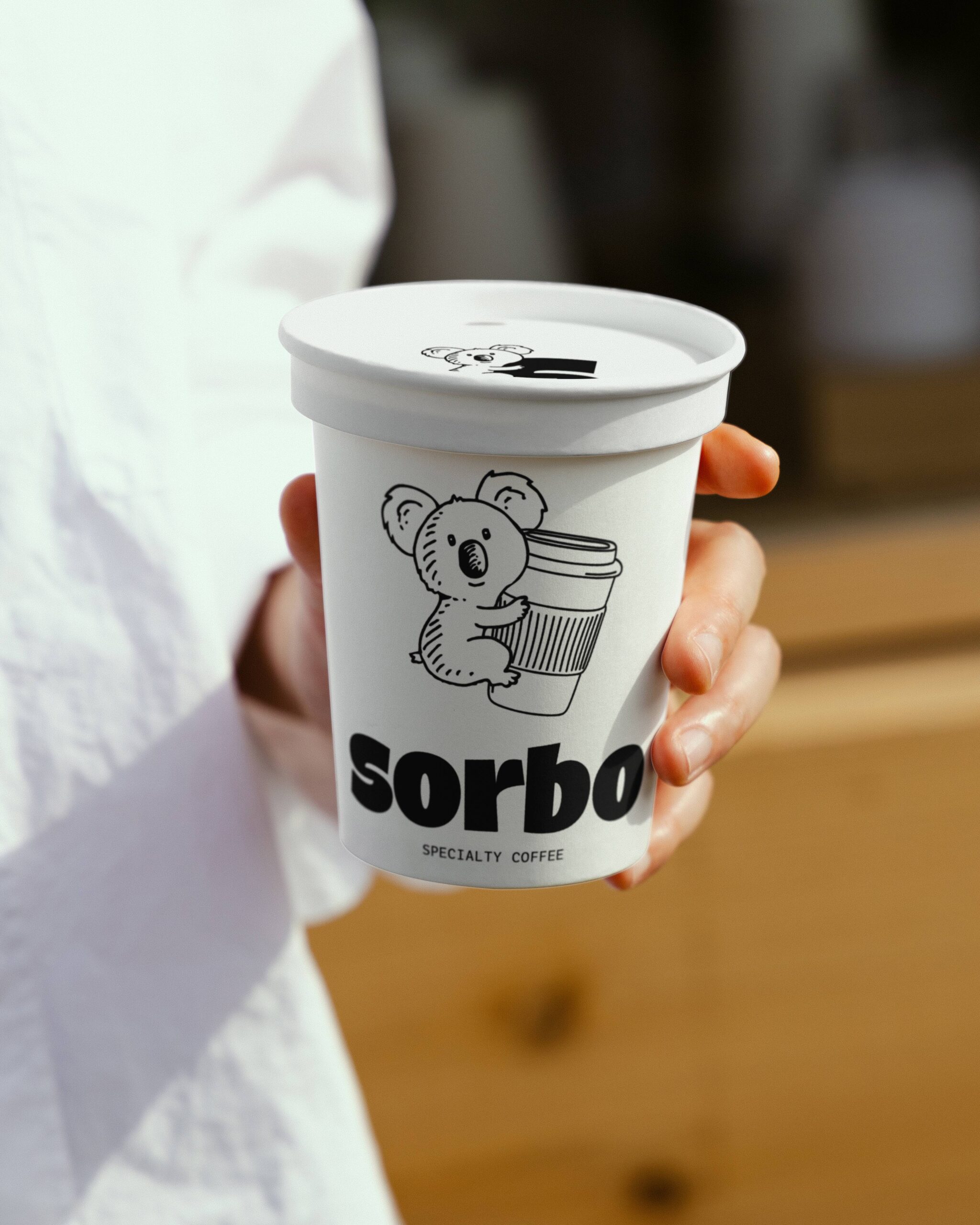Primer plano de un vaso de café Sorbo Specialty Coffee con el logotipo del koala abrazando un vaso, sostenido por una persona.