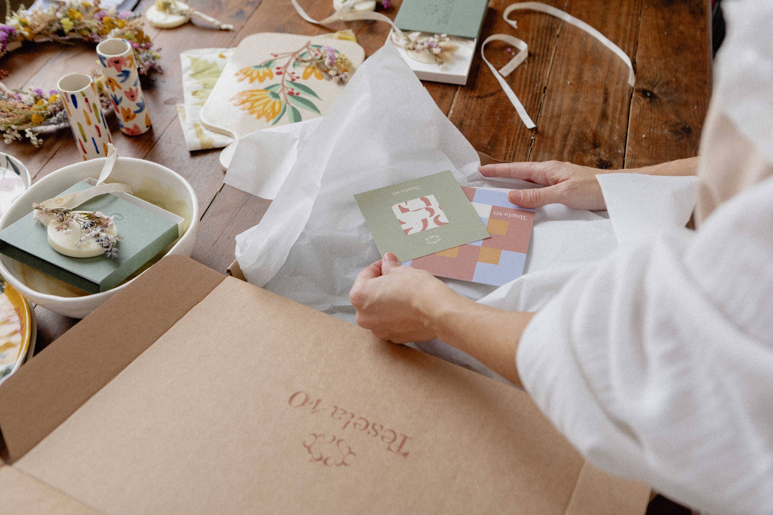 Bodegón de trabajo de branding: caja de envío abierta con papel de seda, tarjetas de marca y elementos decorativos de cerámica sobre mesa de madera.