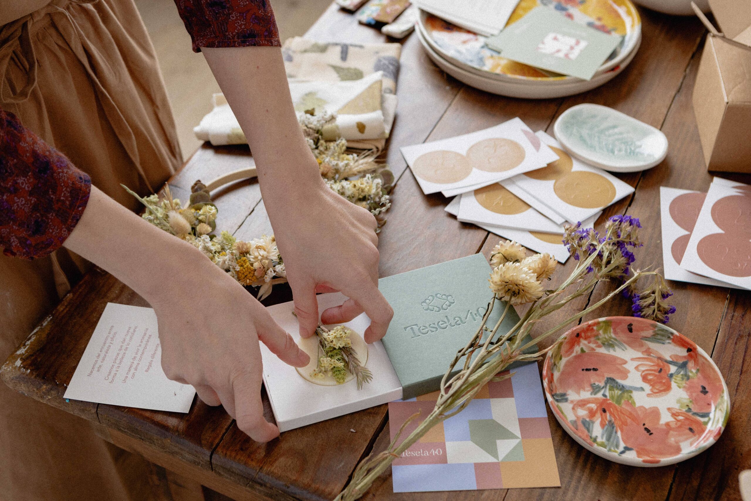 Detalle de packaging experiencial: manos colocando cuidadosamente un pequeño ramillete de flores secas sobre un jabón artesanal en su estuche, junto a una caja verde con logotipo en golpe seco de Tesela 40.