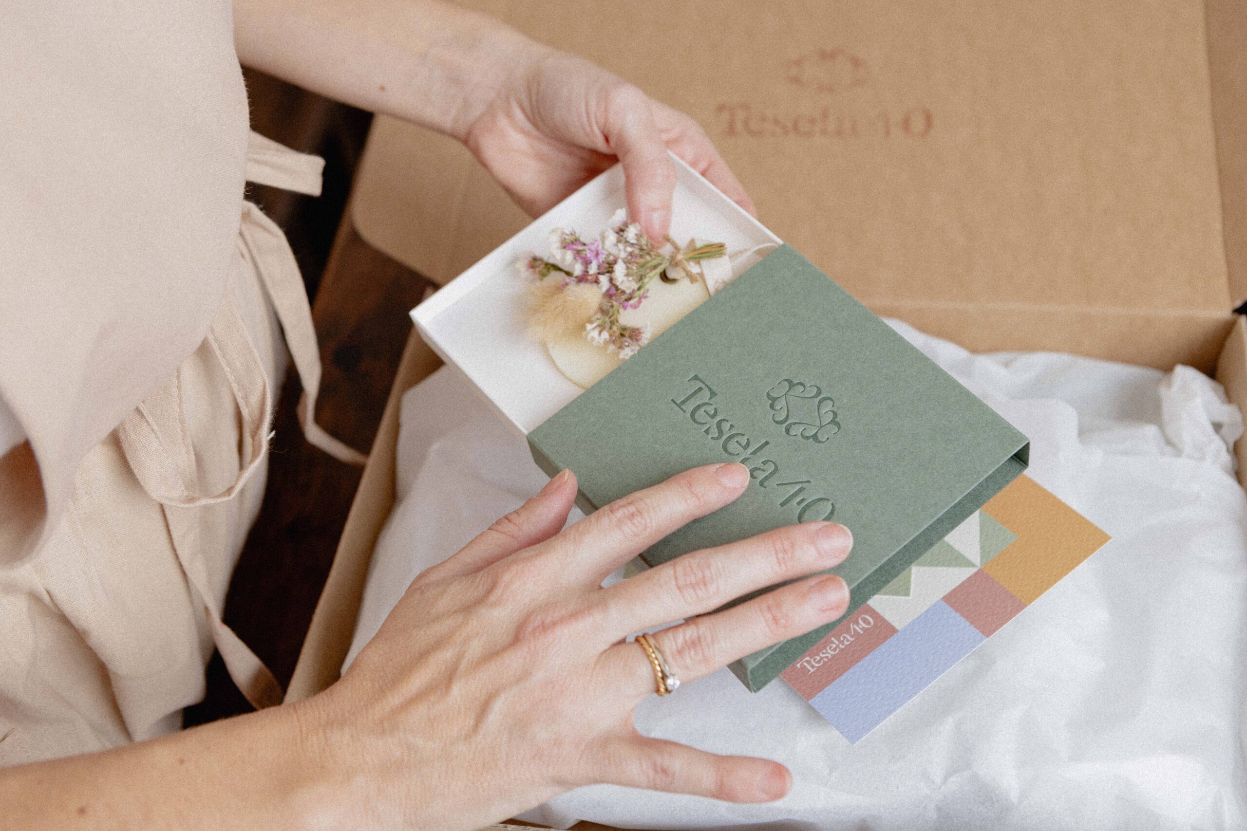 Experiencia de unboxing premium: manos extrayendo un estuche rígido verde oliva con logotipo de Tesela 40 en relieve de una caja de envío protegida con papel de seda