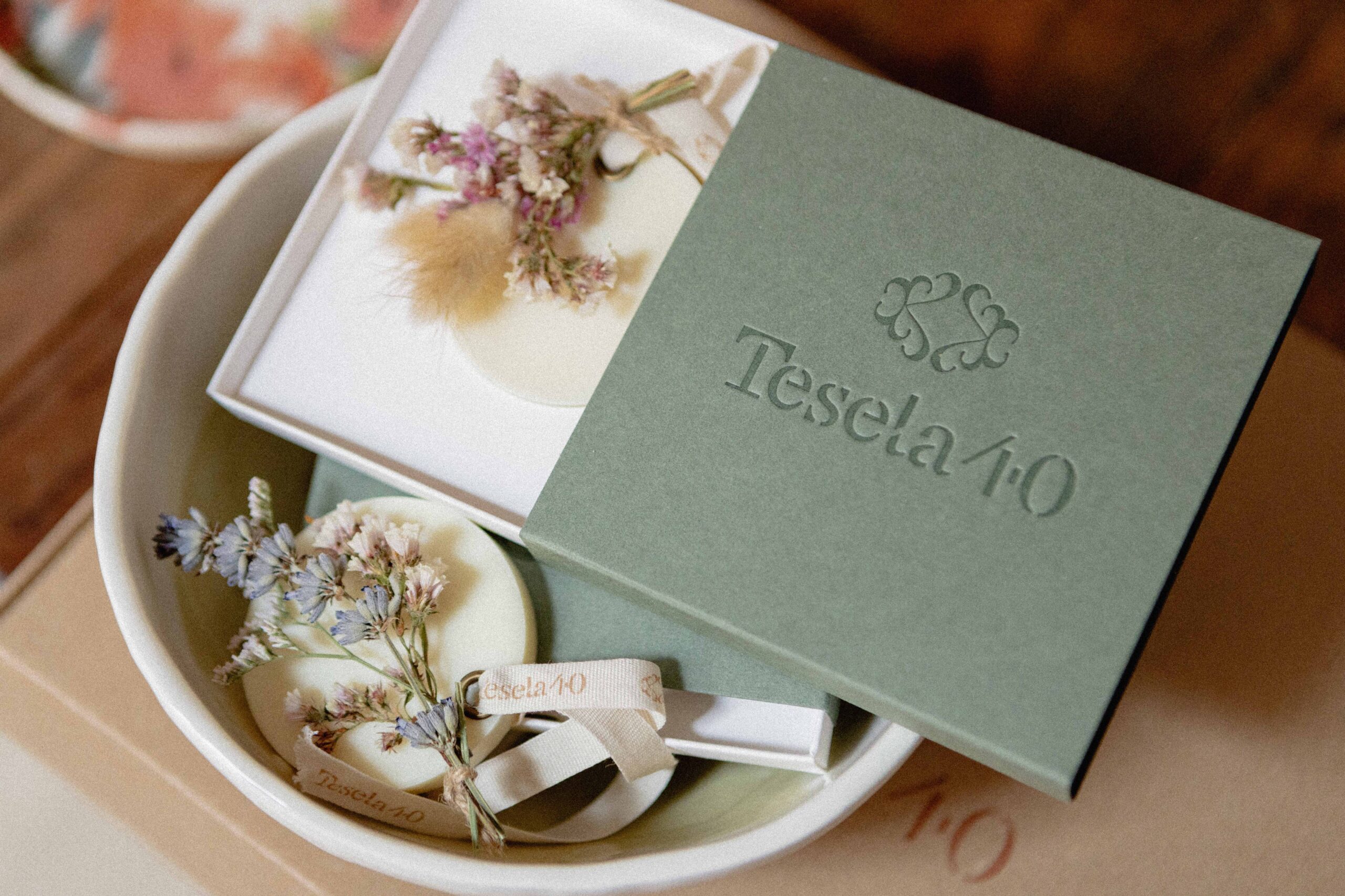 Detalle de packaging premium: caja rígida de color verde oliva con logotipo de Tesela 40 en golpe seco, acompañada de flores secas y ambientador sólido.