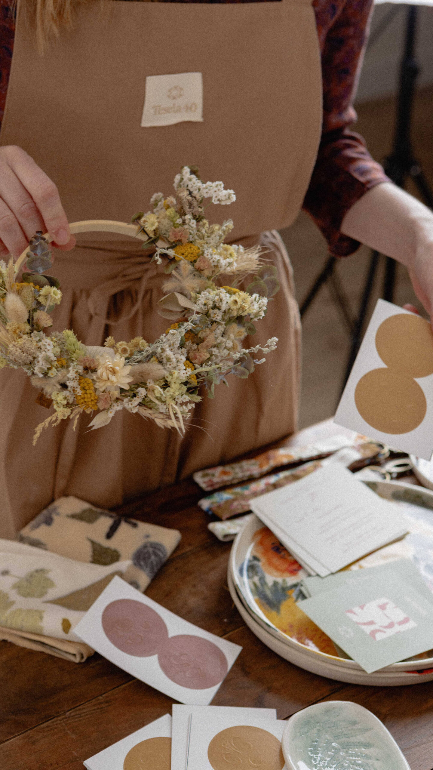 Detalle de artesana con delantal corporativo de Tesela 40 sosteniendo una corona de flores secas, rodeada de elementos de papelería y diseño de marca.